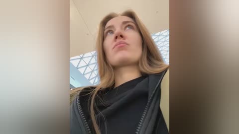 Video of a young Caucasian woman with light skin and long, straight brown hair, wearing a black jacket, looking upward in a modern building with large glass windows and geometric designs.