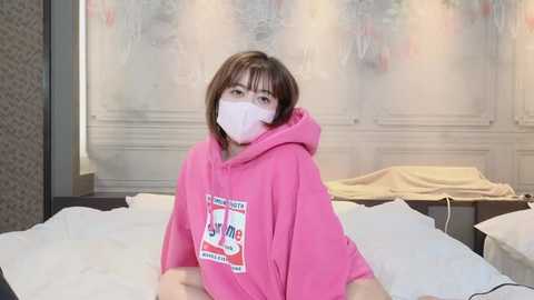 Media: Video of a young East Asian woman with short brown hair, wearing a pink hooded sweatshirt, white surgical mask, and sitting on a bed with white sheets and beige pillows. The background shows a blurred, soft-focus room with light-colored walls.