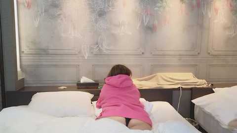 Media: Video of a woman in a pink robe, kneeling on a white bed with a beige blanket, facing a gray, textured wall with abstract art.