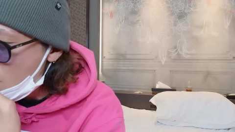 Media: Video of a woman in a pink hoodie, gray beanie, and face mask, standing indoors near a glass door with frosty condensation. Background shows a white pillow and scattered objects on a dark table.