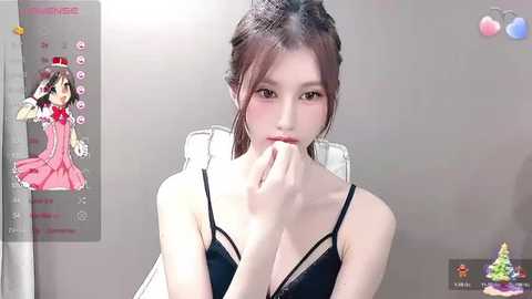 Video of a young East Asian woman with fair skin, brown hair in a bun, wearing a black bra. Background includes a virtual avatar in a pink dress and a digital camera.