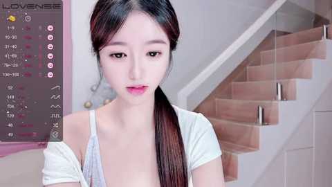 Media: Video of an East Asian woman with fair skin, long black hair, and pink lipstick, wearing a white off-shoulder top. Background features a modern staircase and a digital calendar.