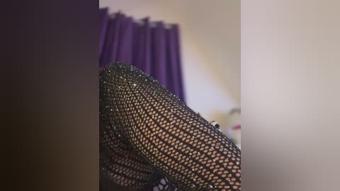 Media: Video of a close-up of a person wearing black fishnet stockings, with a blurred purple curtain in the background.