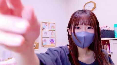 Media: Video of an East Asian woman with long brown hair, wearing a blue mask and dark shirt, holding a hand sanitizer bottle, in a brightly lit room with framed pictures on the wall and a clock.