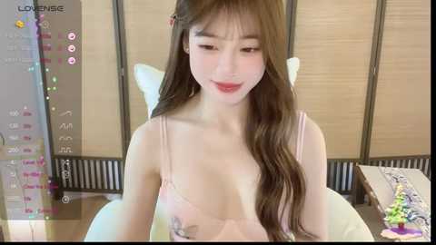 Media: A video of an East Asian woman with long brown hair, fair skin, and light makeup, wearing a pink lingerie top, sitting indoors, looking downward.