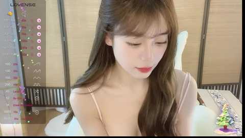 Media: A video of an East Asian woman with long, straight brown hair, wearing a light pink spaghetti-strap top, seated at a table. She has fair skin and closed eyes, looking down. The background features a beige curtain and a partially visible table with a colorful toy.