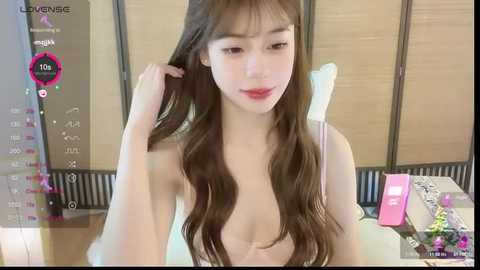 Video of a young Asian woman with long, wavy brown hair, fair skin, and red lipstick, sitting in a modern room with a pink phone and a white chair.