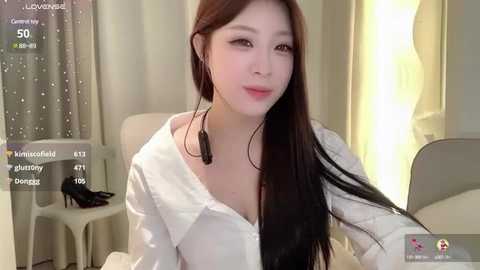 Media: Video of a young Asian woman with long, straight brown hair, wearing a white blouse, seated in a modern, softly lit room.