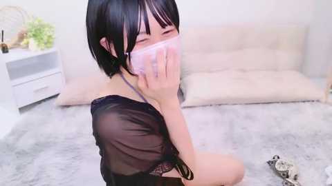 Media: A video of a young Asian woman with short black hair, wearing a sheer black top and a pink face mask, kneeling on a light gray rug in a minimalist, brightly lit room with a white couch and plant in the background.