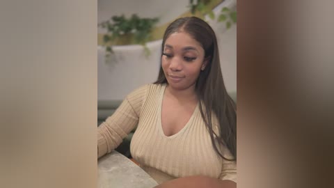 Media: Video of a young Black woman with long, straight brown hair, wearing a cream ribbed sweater, smiling softly. Background features a blurred indoor setting with green plants.