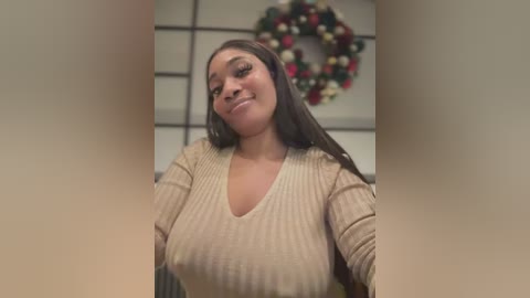 Media: A video of a smiling, light-skinned Black woman with long, straight dark hair, wearing a beige ribbed V-neck sweater, standing indoors near a festive wreath on a wall.