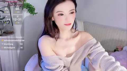 Media: Video of a fair-skinned, young Asian woman with long black hair, wearing a beige off-shoulder sweater, seated on a bed with a white headboard.