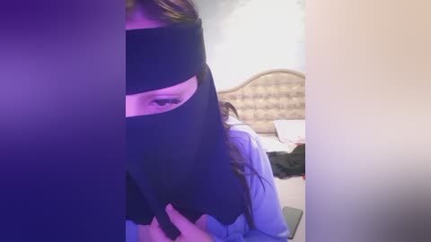 Media: Video of a person with a black mask covering the eyes, sitting in a dimly lit bedroom with a white bed and headboard visible in the background.