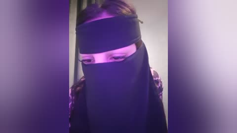 Media: A video of a person with brown hair wearing a black headband, standing in a dimly lit room with purple lighting. The person's face is partially covered by a black veil.