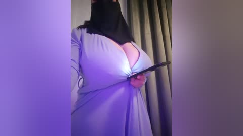 Media: Video of a woman with a large bust in a light gray dress, wearing a black hijab and holding a black phone, standing in a dimly lit room with green curtains.