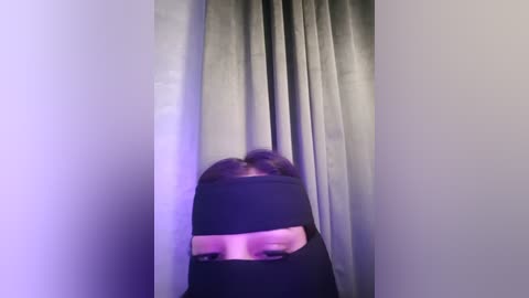 Media: A blurry video of a person with short, dark hair partially obscured by a black cloth, standing in front of light gray curtains in a dimly lit room.