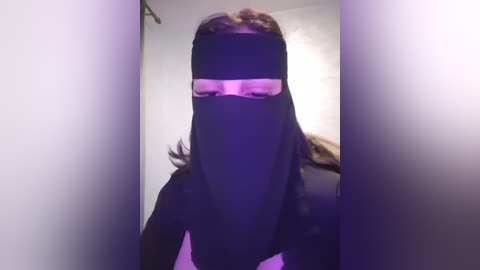 Media: Video of a person wearing a black face mask, head tilted slightly, standing in a dimly lit room with a purple tint.