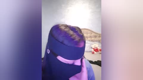 Media: Video of a person wearing a black face mask and purple hair, standing in a bedroom with a bed, stuffed animal, and wall with a light color.