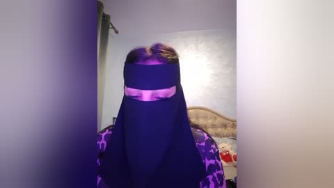 Media: Video of a person in a black burqa with a pink belt, sitting on a bed with a patterned blanket in a dimly lit, plain-walled bedroom.