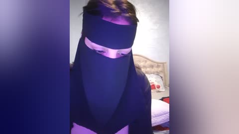 Media: A video of a person wearing a black niqab with a white band, standing in a bedroom with a beige bed, pillows, and a floral-patterned blanket. The room is dimly lit.