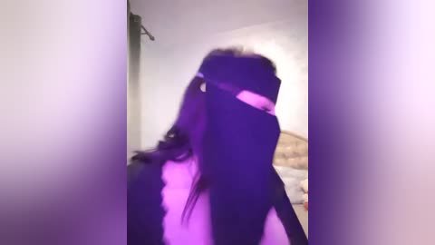 Media: A video of a person with long, wavy hair, wearing a black shirt, standing in a dimly lit room with purple lighting, creating a moody, atmospheric effect.