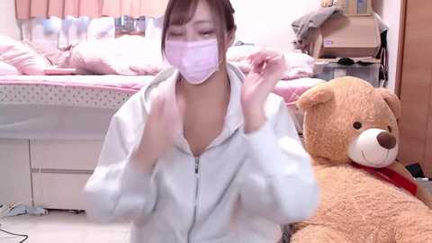 Media: Video of an Asian woman in a light pink face mask, white hoodie, adjusting her mask, surrounded by a messy bedroom with a bed, teddy bear, and clutter.