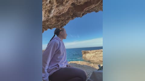 Media: Video of a young woman with a fair complexion and long, dark hair tied back, wearing a light purple shirt and dark pants, seated on a rocky shore under a natural cave arch, gazing out at a clear blue sky and ocean.