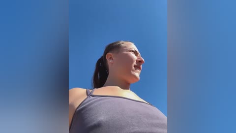 Media: Video of a smiling woman with light skin and brown hair in a ponytail, wearing a gray tank top, standing against a clear blue sky.