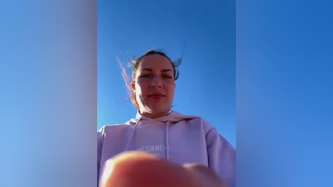 Media: Video of a young woman with fair skin and dark hair, wearing a light purple hoodie, viewed from a low angle against a clear blue sky.