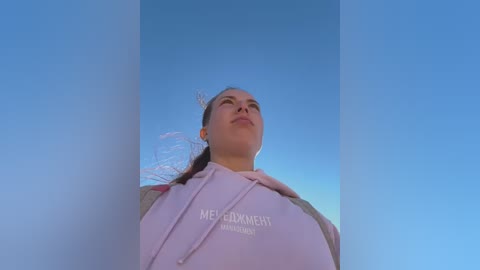 Media: A video of a young woman with light skin and brown hair, wearing a light pink hoodie, standing against a clear blue sky.