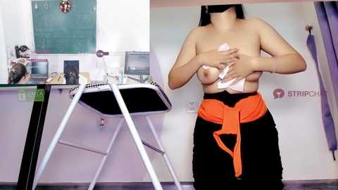 Media: Video of a topless woman with medium-sized breasts, wearing a black skirt and orange belt, in a classroom setting with a desk, chair, and chalkboard.