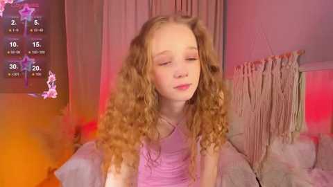 Media: Video of a young girl with curly blonde hair, wearing a light pink dress, sitting on a bed with pink and beige bedding, surrounded by warm, glowing pink and orange lighting.