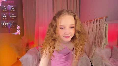 Media: Video of a young girl with curly, light brown hair, wearing a purple top, smiling in a dimly lit room with a pinkish hue.