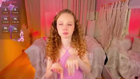 Media: Video of a young, fair-skinned girl with curly brown hair, wearing a purple halter top, seated on a beige couch under pink and orange lighting.