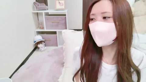 Media: Video of an East Asian woman with long brown hair, wearing a white face mask, white top, and sitting on a white couch in a pastel-themed, minimalist room with white shelves and pink storage bins.