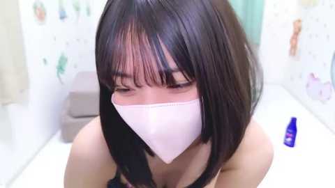 Media: A video of a young Asian woman with straight black hair and bangs, wearing a white face mask, standing in a white-tiled bathroom with a blue bottle on the sink.