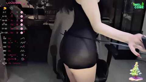 Media: Video of a woman in a sheer black bodysuit, showcasing her bare back and buttocks. Background shows a dimly lit room with a TV screen displaying a camera feed.