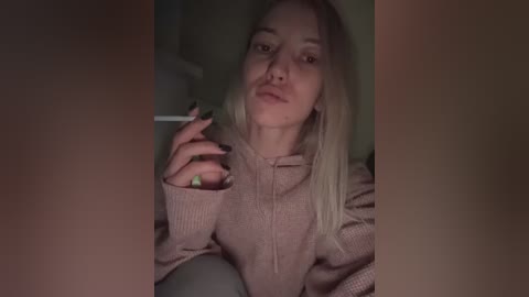 Media: Video of a blonde woman with light skin, wearing a light pink hoodie, holding a cigarette, looking into a phone, dimly lit background.