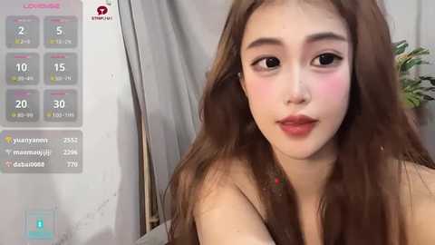 Media: Video of an Asian woman with long brown hair, light skin, and full lips, wearing red lipstick, looking at the camera. Background shows a white wall with a digital thermometer displaying 20 degrees Celsius.