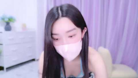 Media: Video of an Asian woman with long black hair, wearing a white mask, blue top, and sitting on a beige chair in a pastel-colored room.