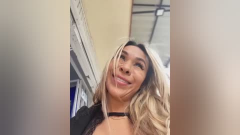 Media: Video of a smiling Latina woman with long, wavy blonde hair, wearing a black lace top and choker necklace, captured from a low angle.