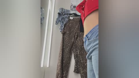 Media: Video of a woman's lower torso in a dressing room. She wears a red crop top and blue jeans, with leopard-print pajamas and a plaid shirt hanging nearby.