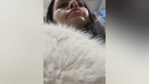 Media: A video captures a close-up, upward-facing shot of a woman's face and fluffy white fur, likely a pet. The background is blurred, with blue and white tones. The woman has long, dark hair and light skin.