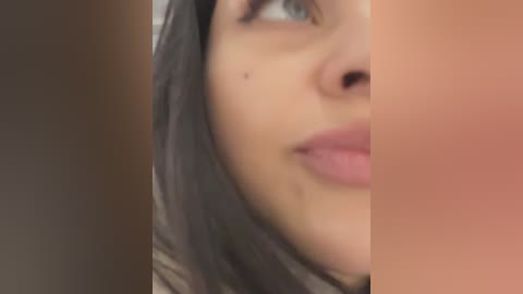 Media: A close-up video of a woman's face, focusing on her left eye, nose, and lips. Her skin is light with a mole on her cheek, and she has straight, dark brown hair. The background is blurry, and her expression is neutral.