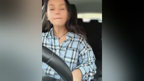 Media: A video of a woman with long dark hair, wearing a plaid shirt, driving a car with a black seatbelt across her chest.