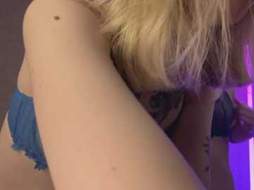 Media: Video of a blonde woman with fair skin, wearing blue panties, leaning forward with her breasts visible. The background is dark with purple lighting.