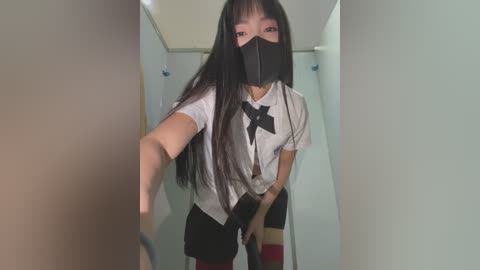 Media: A video of an East Asian woman with long black hair, wearing a white blouse, black bow tie, black mask, and black shorts, taking a selfie in a narrow, beige hallway.