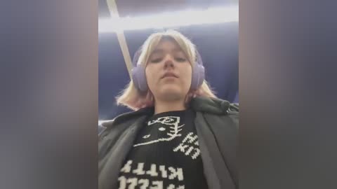 Media: Video of a woman with short blonde hair, wearing purple headphones, a black T-shirt with white text, and a dark jacket. She appears relaxed in a dimly lit, possibly underground train.