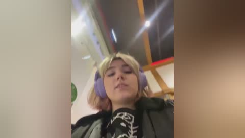 Media: Video of a young woman with blonde hair, wearing purple headphones and a black graphic tee, standing indoors against a blurred background.