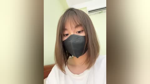 Media: Video of an East Asian woman with straight, shoulder-length brown hair, wearing a black face mask and a white t-shirt, standing in a minimalist room with pale green walls and an air conditioning unit.